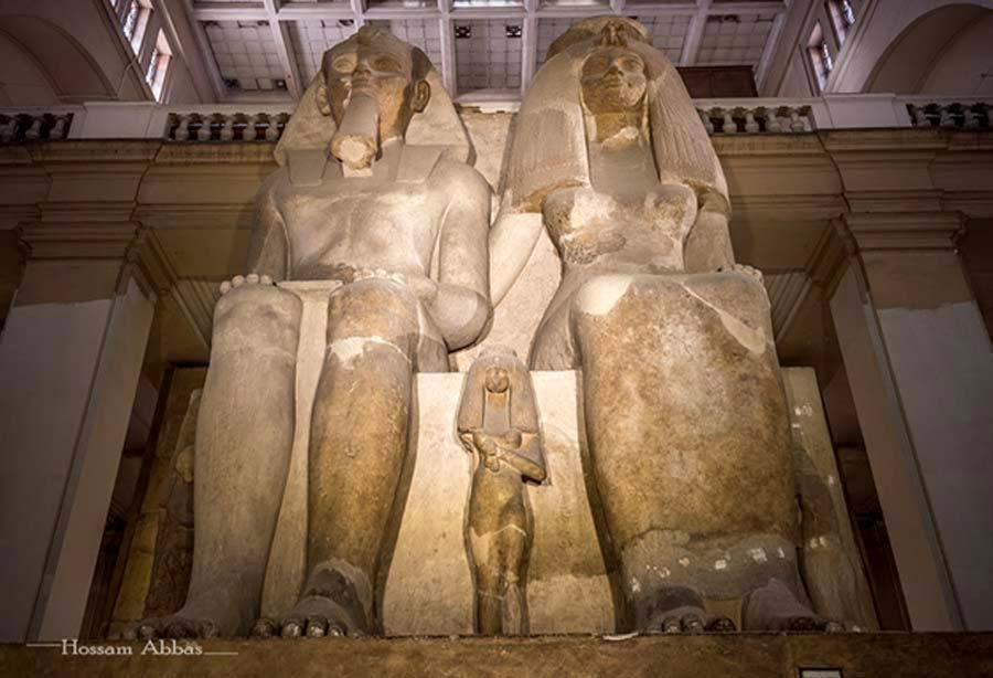 The colossal limestone group statue of Amenhotep III, Queen Tiye and their daughters – Henuttaneb, the largest and best preserved, in the center; Nebetah on the right; and another, whose name is destroyed. The sculpture rises seven meters in height dwarfing all other statues in the central hall of the Egyptian Museum, Cairo. This monolith originated from Medinet Habu in Western Thebes.